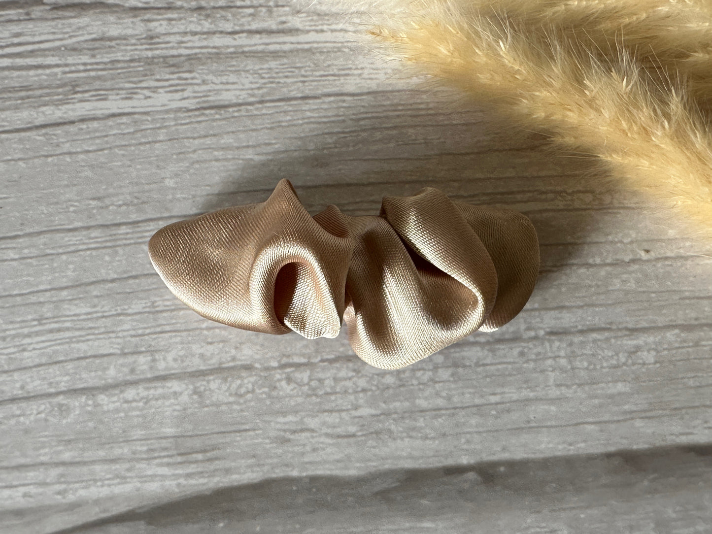 The Champagne Gold Silk French Barrette Scrunchie Clip by Crafts by Kate exudes luxury on a light wooden surface, complemented by the charm of a fluffy yellow accent in the corner.