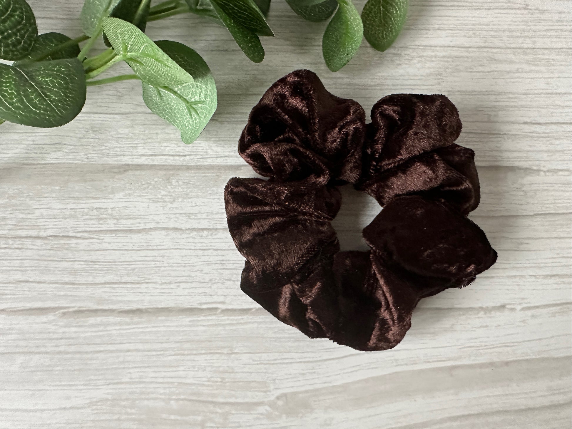 The Crafts by Kate Crushed Velvet Extra Large Scrunchie in Chocolate Brown rests on a light wood surface beside green artificial leaves.