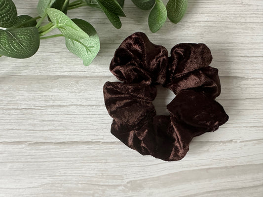 The Crafts by Kate Crushed Velvet Extra Large Scrunchie in Chocolate Brown rests on a light wood surface beside green artificial leaves.
