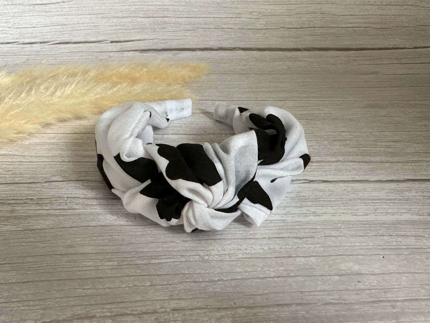 A Cotton Knotted Headband in black cow print by Crafts by Kate sits on a light wood surface beside a beige pampas grass stem.