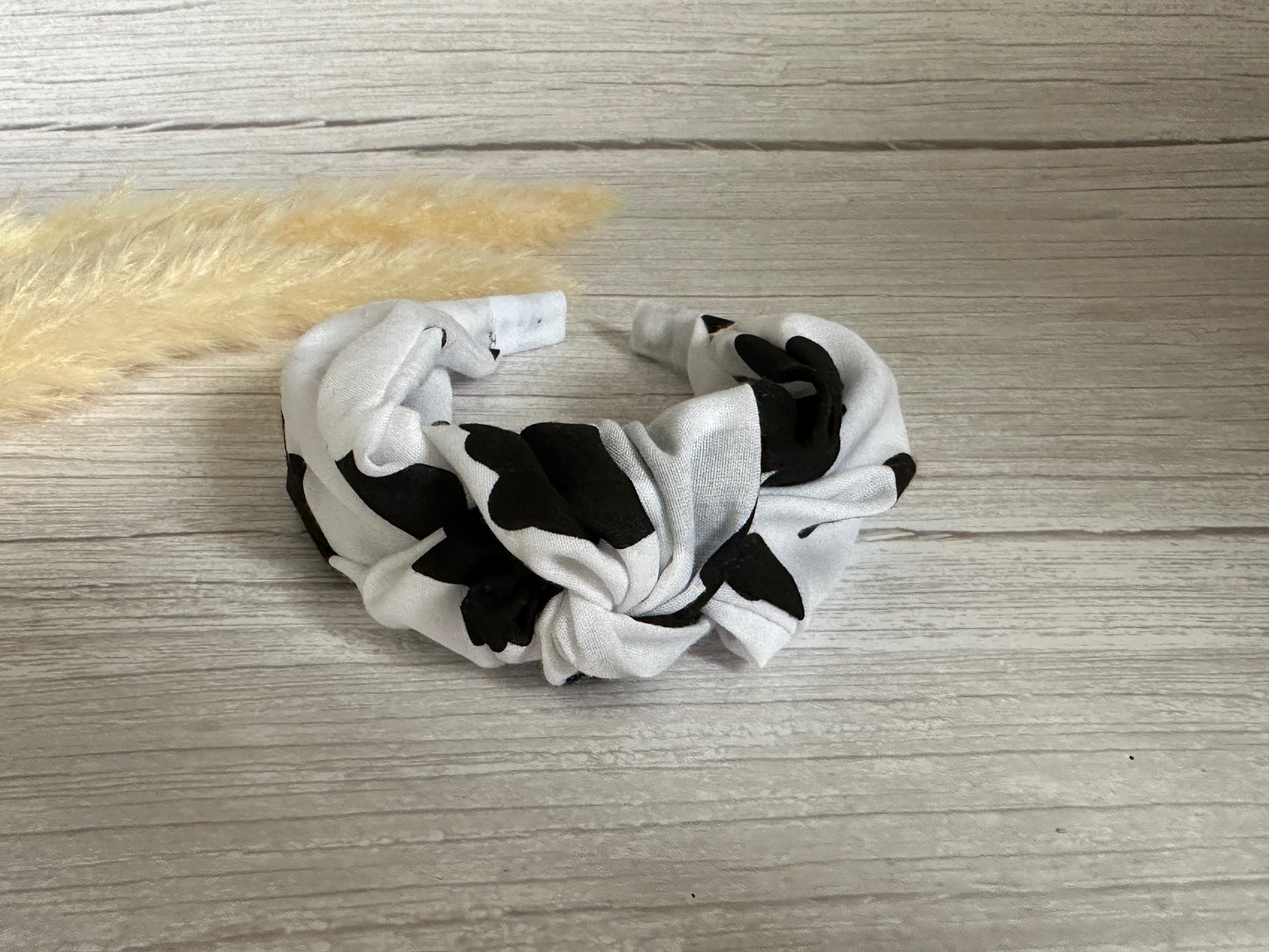 A Cotton Knotted Headband in black cow print by Crafts by Kate sits on a light wood surface beside a beige pampas grass stem.