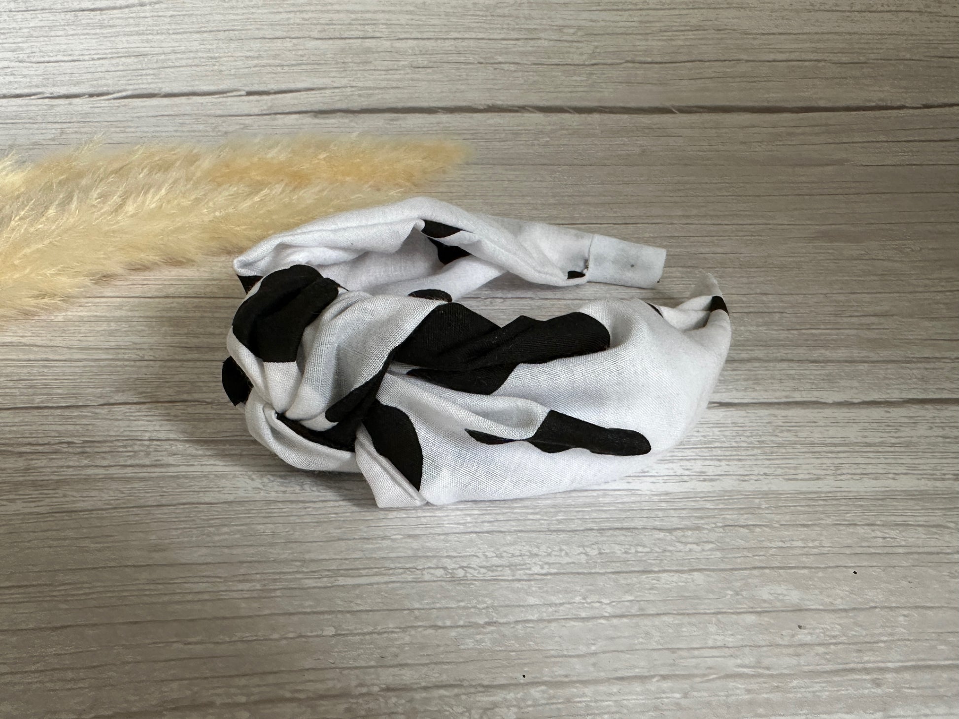 The Cotton Knotted Headband - Cow Print by Crafts by Kate rests on a light wood surface beside beige pampas grass.