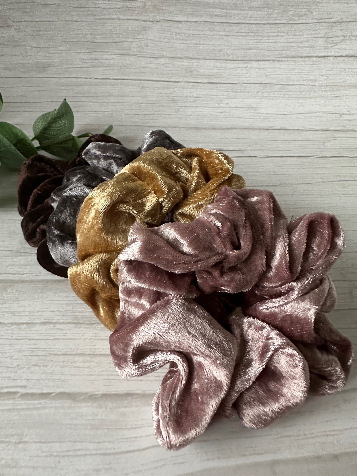A close-up of five Crafts by Kate Crushed Velvet Extra Large Scrunchies in luxury gold, brown, and blush pink, stacked on a light wooden surface with a green leafy plant in the background.