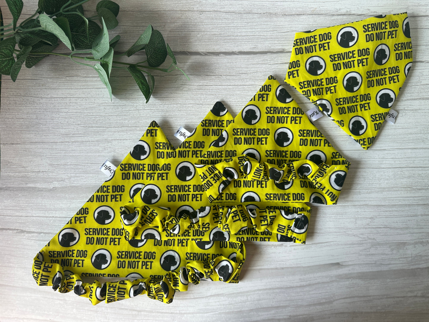 The Cotton Pet Scrunchie Bandana - Anxious by Crafts by Kate features a yellow handcrafted bandana with black "Service Dog Do Not Pet" text and paw prints, plus cotton scrunchies, shown on light wood beside a leafy green plant.