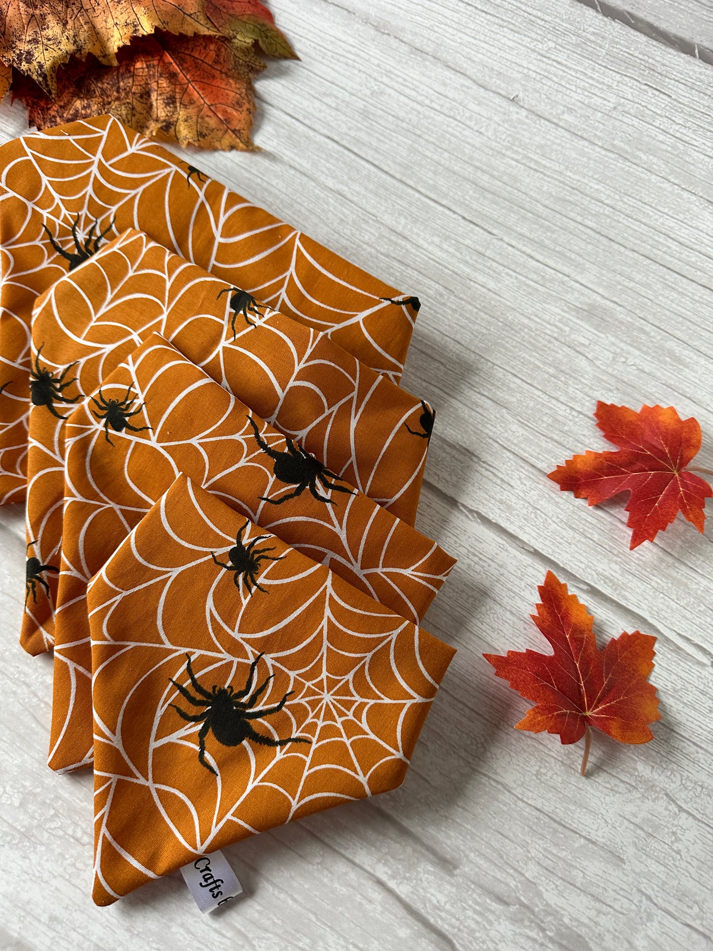 Five orange cloth napkins featuring a white spiderweb and black spider design are fan-folded on a light-colored wooden surface. Red and orange autumn leaves are scattered around the napkins, adding a festive, fall-themed touch with a hint of Crafts by Kate's Cotton Pet Scrunchie Bandana - Spider Vibes in the setting.