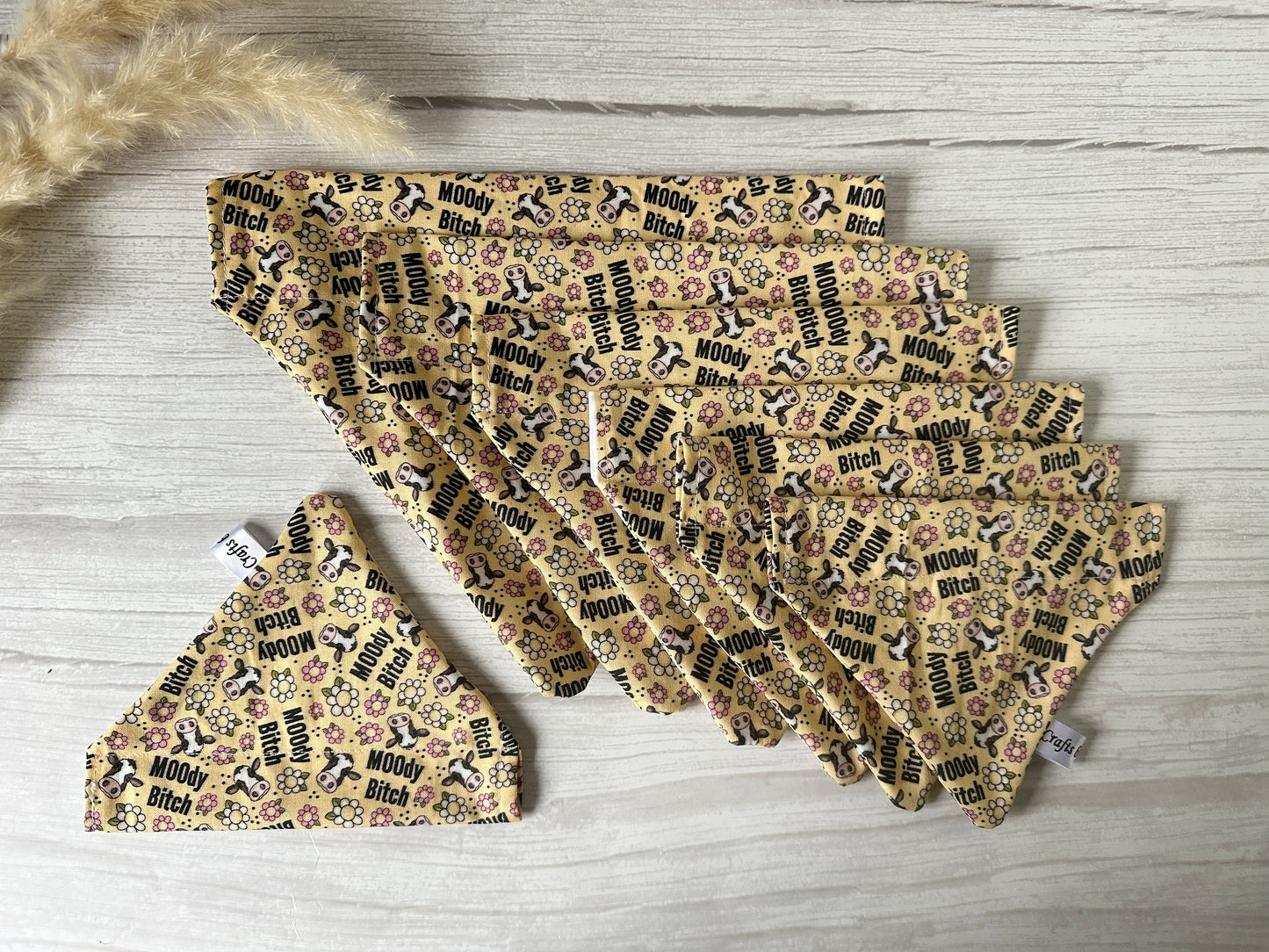 A collection of Crafts by Kate's Cotton Pet Bandanas with collar attachment, labeled "Moody Cow," are neatly displayed on a wooden surface. These cream bandanas feature "Moody Bitch" text along with small pink florals, while dried grass is visible in the corner.