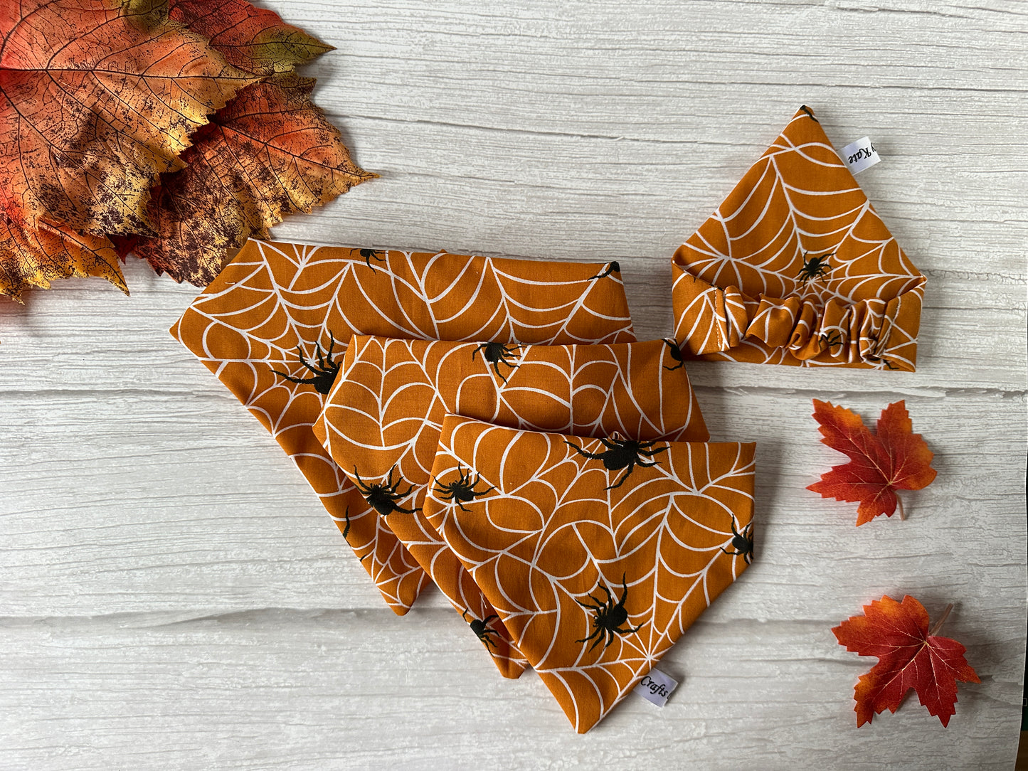 Four pieces of the "Cotton Pet Scrunchie Bandana - Spider Vibes" from Crafts by Kate, featuring orange fabric with white spiderweb patterns and small black spiders, are displayed on a white wooden background. The items include a pet bandana, bow tie, Handcrafted Cotton Pet Scrunchie, and a bow. Autumn leaves are scattered around them.
