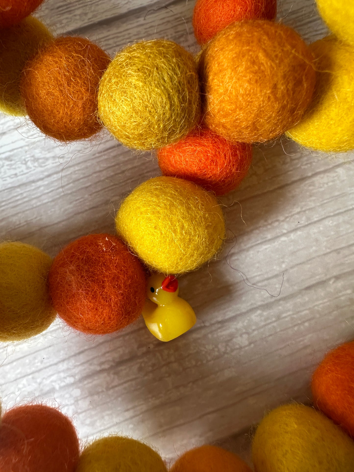 Close-up of the PomPom Pet Collar - Rubber Ducks by Crafts by Kate, featuring yellow and orange felt balls on an elastic string and a small yellow duck charm, displayed on a light wooden surface.