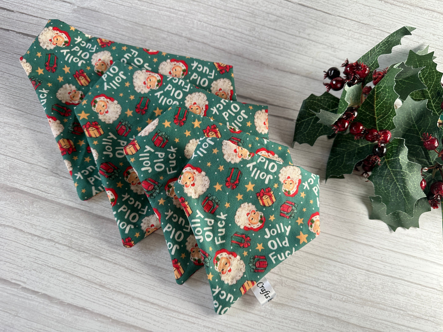 Crafts by Kate's "Cotton Pet Scrunchie Bandana - Jolly Old F***" is showcased on a light wood surface with green festive fabric featuring Santa and gift boxes, alongside red berries and holly. Ideal for crafting a unique pet accessory.