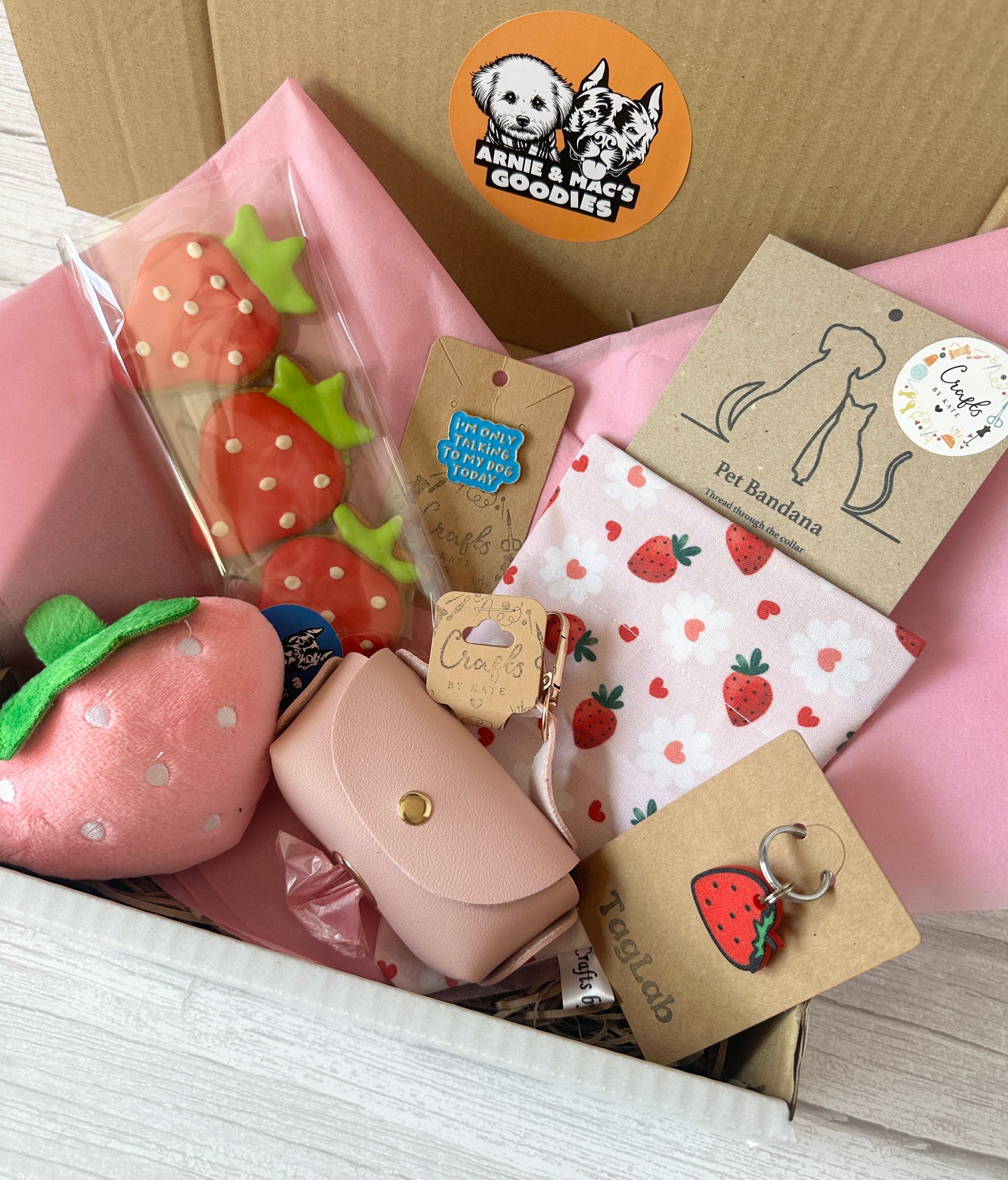 Crafts by Kate’s Strawberries & Daisies - Arnie & Mac’s Goodies gift box includes strawberry-themed dog cookies, peanut butter treats, a plush toy, bandana, pink treat pouch, keychain, and more—beautifully arranged on soft pink tissue paper.