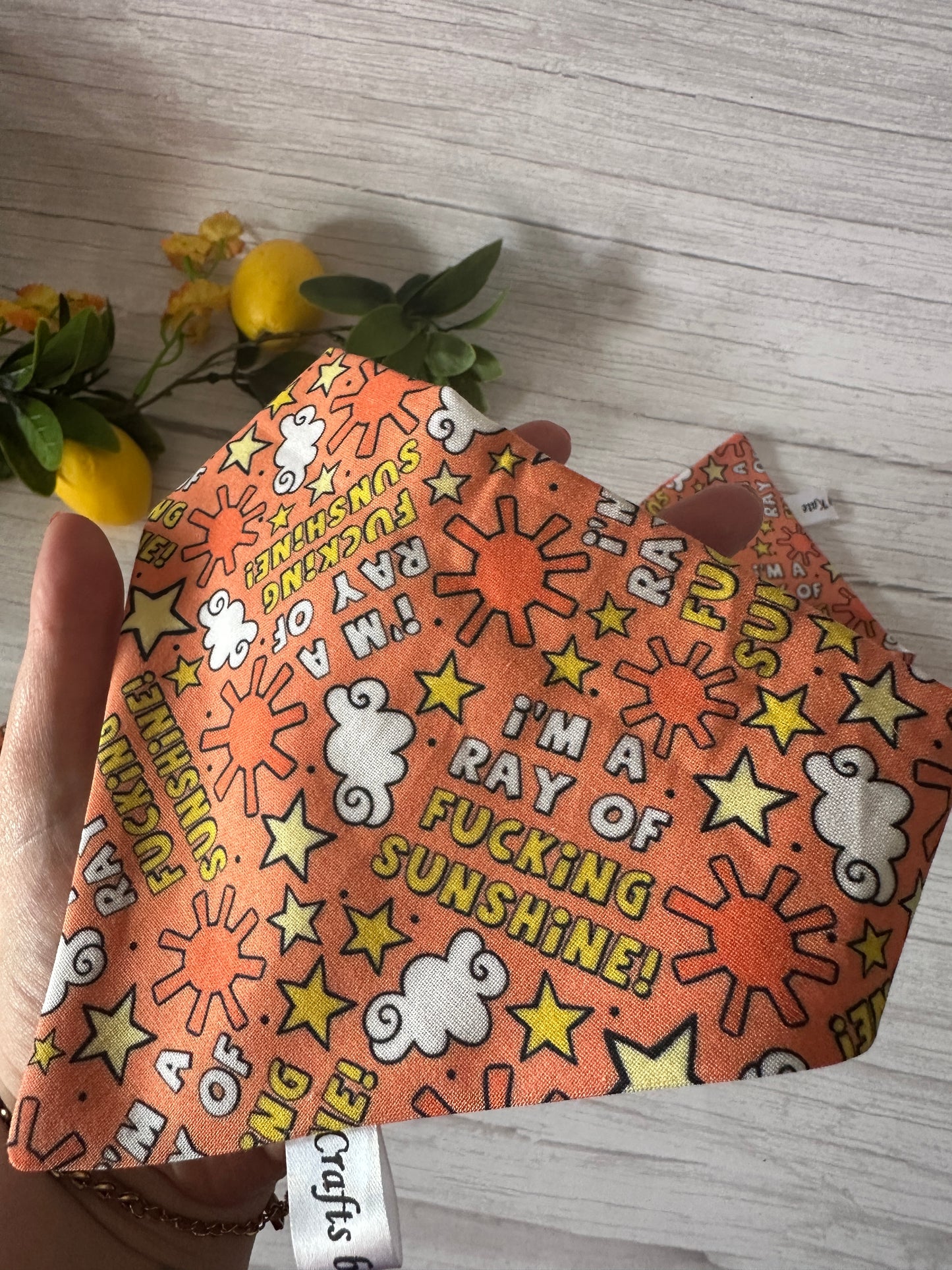 A hand holds an orange fabric mask with suns, stars, and “I’m a ray of f***ing sunshine!” Lemons and yellow flowers surround a Crafts by Kate Cotton Pet Scrunchie Bandana on a light wooden surface.