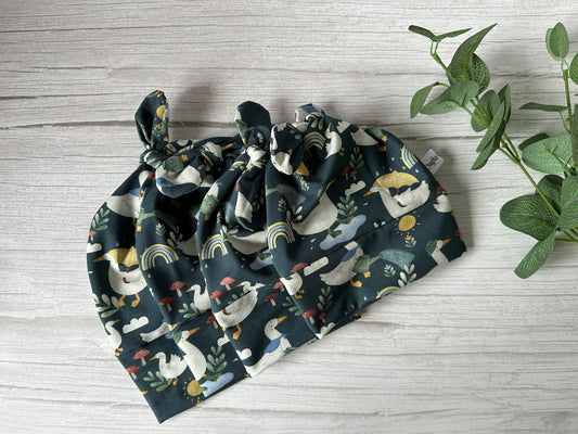 The Baby Wool Cotton Jersey Knotted Hat by Crafts by Kate, featuring sloths, rainbows, and foliage on rainy ducks fabric, is stylishly displayed on a light wood surface. A small leafy plant adds a touch of nature to the right of the wrap.