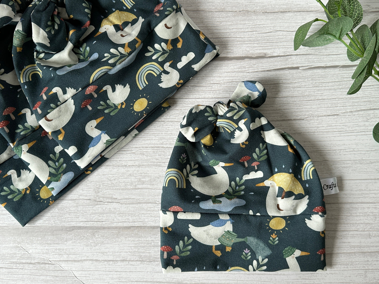 The Baby Wool Cotton Jersey Knotted Hat - Rainy Ducks by Crafts by Kate features a dark green design with duck and mushroom patterns. This cozy luxury wool jersey item adds warmth to rainy days, perfect on a wooden surface beside greenery.