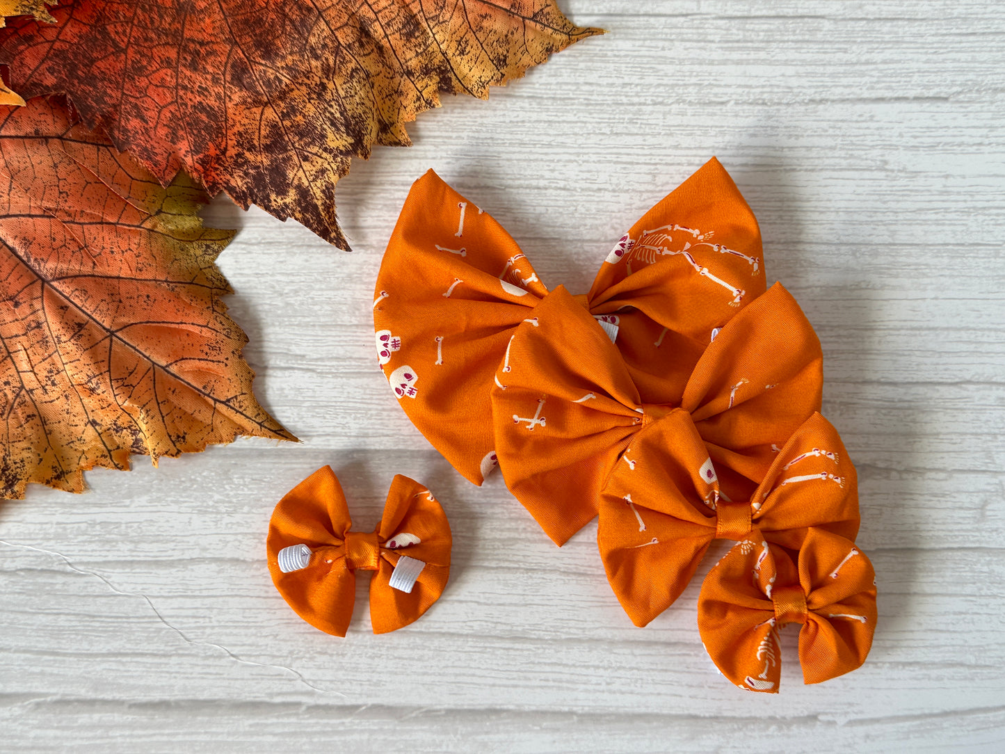Four Crafts by Kate Cotton Pet Bows with collar attachments in the Dancing Skeletons design are displayed on a light wooden surface, accented with autumn leaves. Each bow features elastic for easy use.