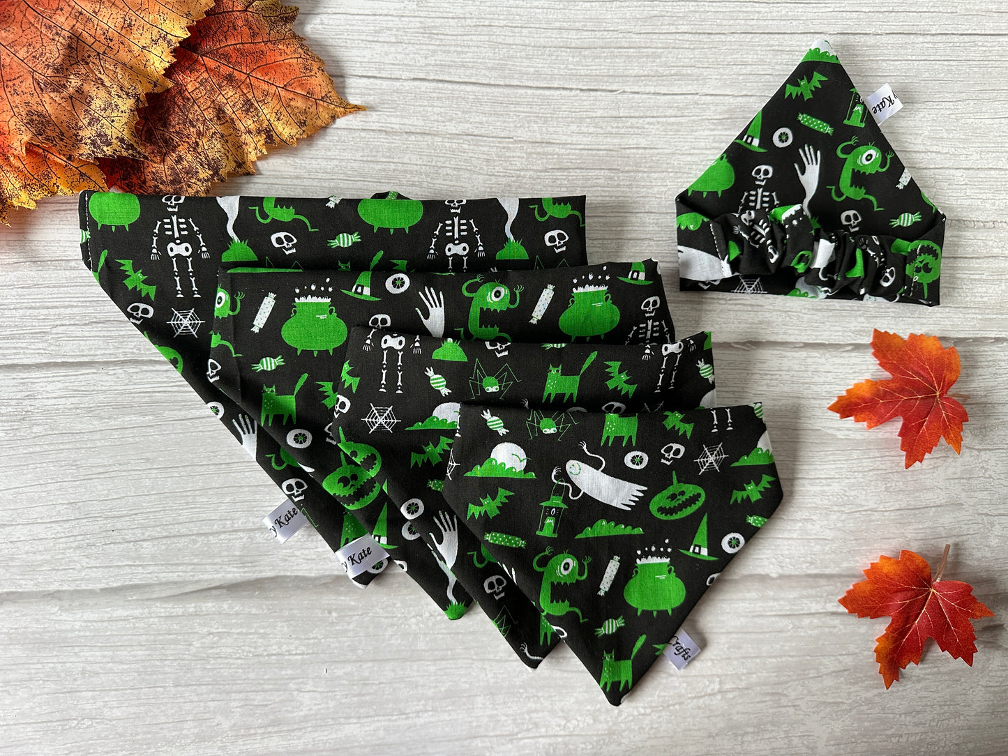 Five black handcrafted “Cotton Pet Scrunchie Bandana - Green Halloween” by Crafts by Kate, featuring green and white ghosts, pumpkins, and cauldrons, are arranged on wood with autumn leaves. Matching bandana and bow tie included.