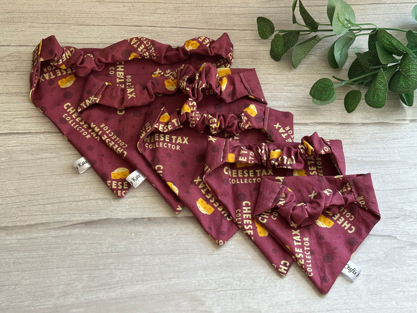 Crafts by Kate's Cotton Pet Scrunchie Bandanas feature maroon-colored Nosé Fabric with a bow tie, available in various sizes. These handcrafted bandanas have a "CHEESE TAX COLLECTOR" print with cheese illustrations, displayed beside greenery on a wooden surface.