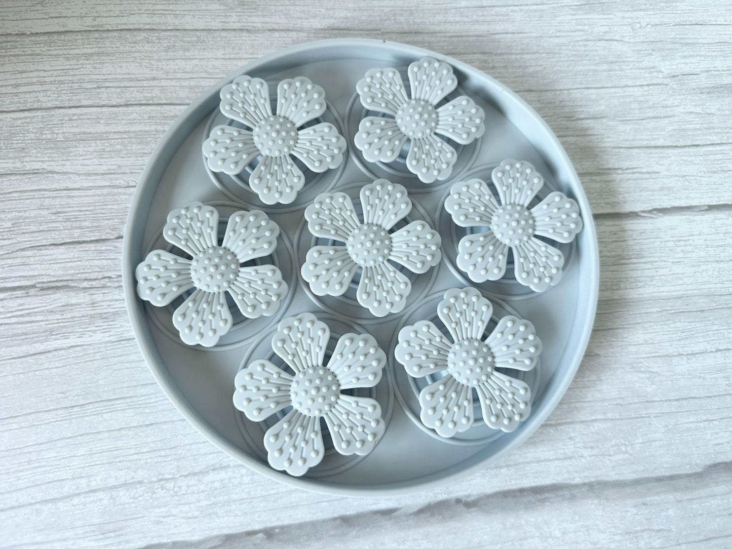 On a light wooden surface, the Crafts by Kate Slow Feeder Pet Mat showcases seven white flower-shaped objects, each with five petals and textured centers that resemble tiny suction cups.