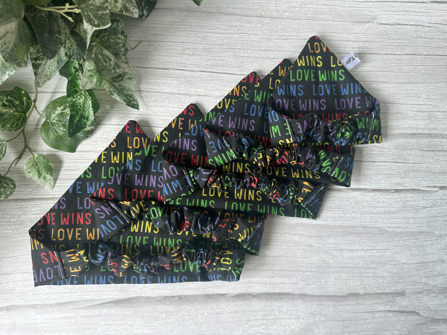 Four black "Love Wins" cotton pet scrunchie bandanas by Crafts by Kate celebrate the LGBTIQA+ community, displayed on light wood with green leafy plants in the top left corner.