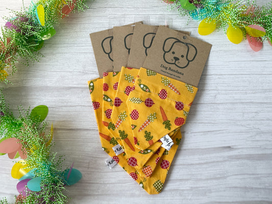Five handcrafted "Vegetable Patch" pet bandanas by Crafts by Kate, made of cotton with fruit and veggie prints, are displayed on a light wood surface. Each slides through the collar and is attached to brown cardboard backing featuring a dog face illustration with colorful decorations.