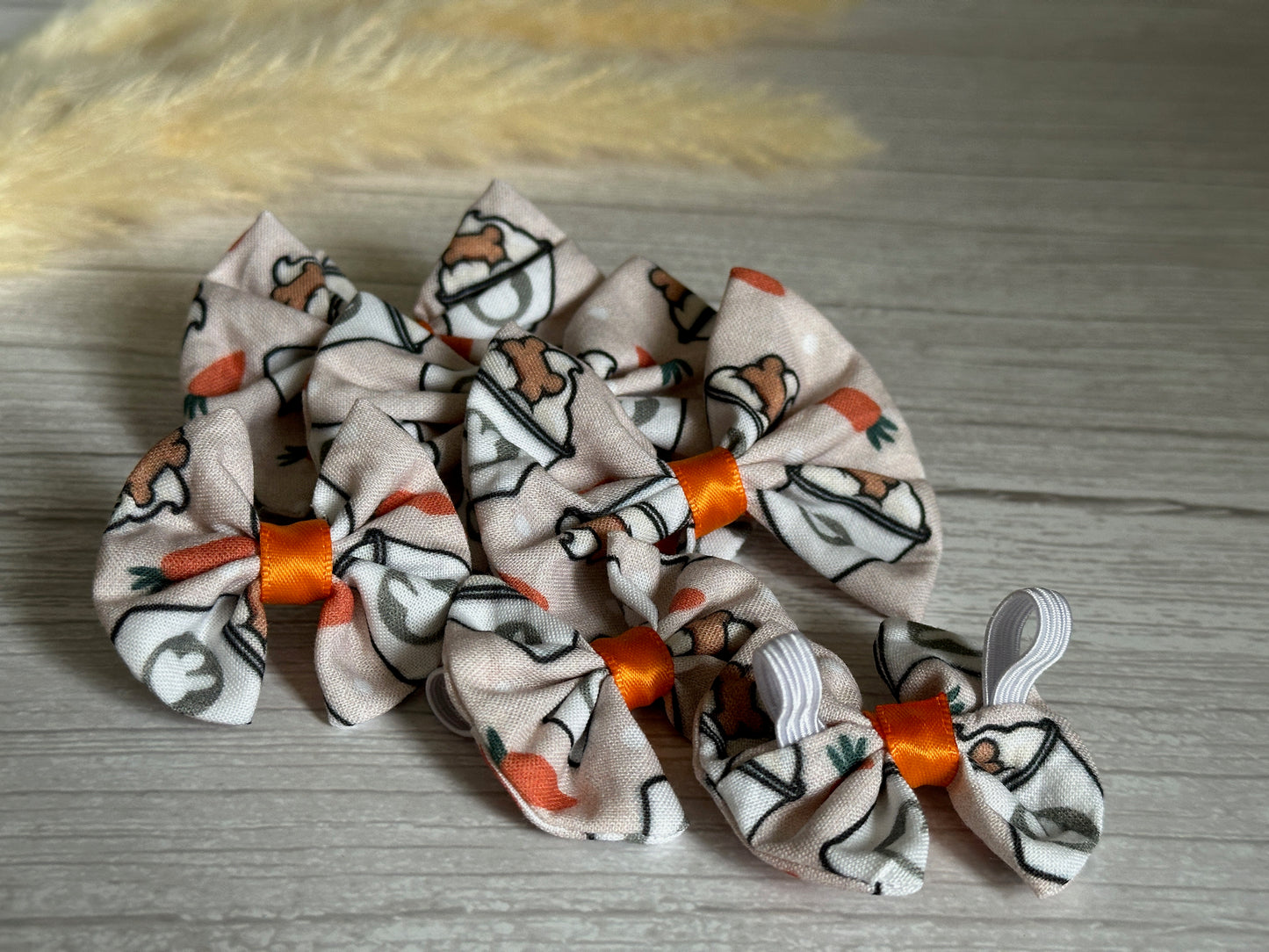 Crafts by Kate's Easter Pupcup collection features cotton pet bows with cartoon bunnies and carrots, each styled with an orange center band. These bows, arranged on wood, come in various sizes and blurred plumes in the background add a dreamy touch.