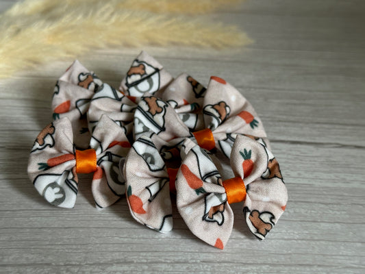 Six Easter Pupcup cotton pet bows by Crafts by Kate, showcasing cartoon rabbits and carrots on a light background with orange center bands, are arranged on a wooden surface. Each bow includes an elastic fastening for easy attachment. A blurred beige plant softly decorates the top left corner.