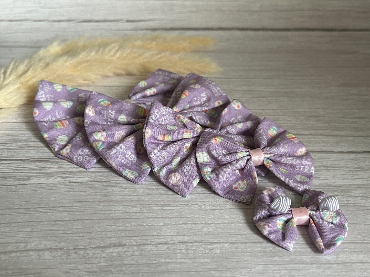 An arrangement of Easter-themed pastel purple bows made from Egg-stra Print Fabric by Crafts by Kate is displayed on a light wood surface with a soft fluffy accent in the back. These handcrafted Cotton Pet Bows feature decorative Easter eggs and printed words like "egg," "straw," and "little," and come with elastic fastenings for easy attachment.