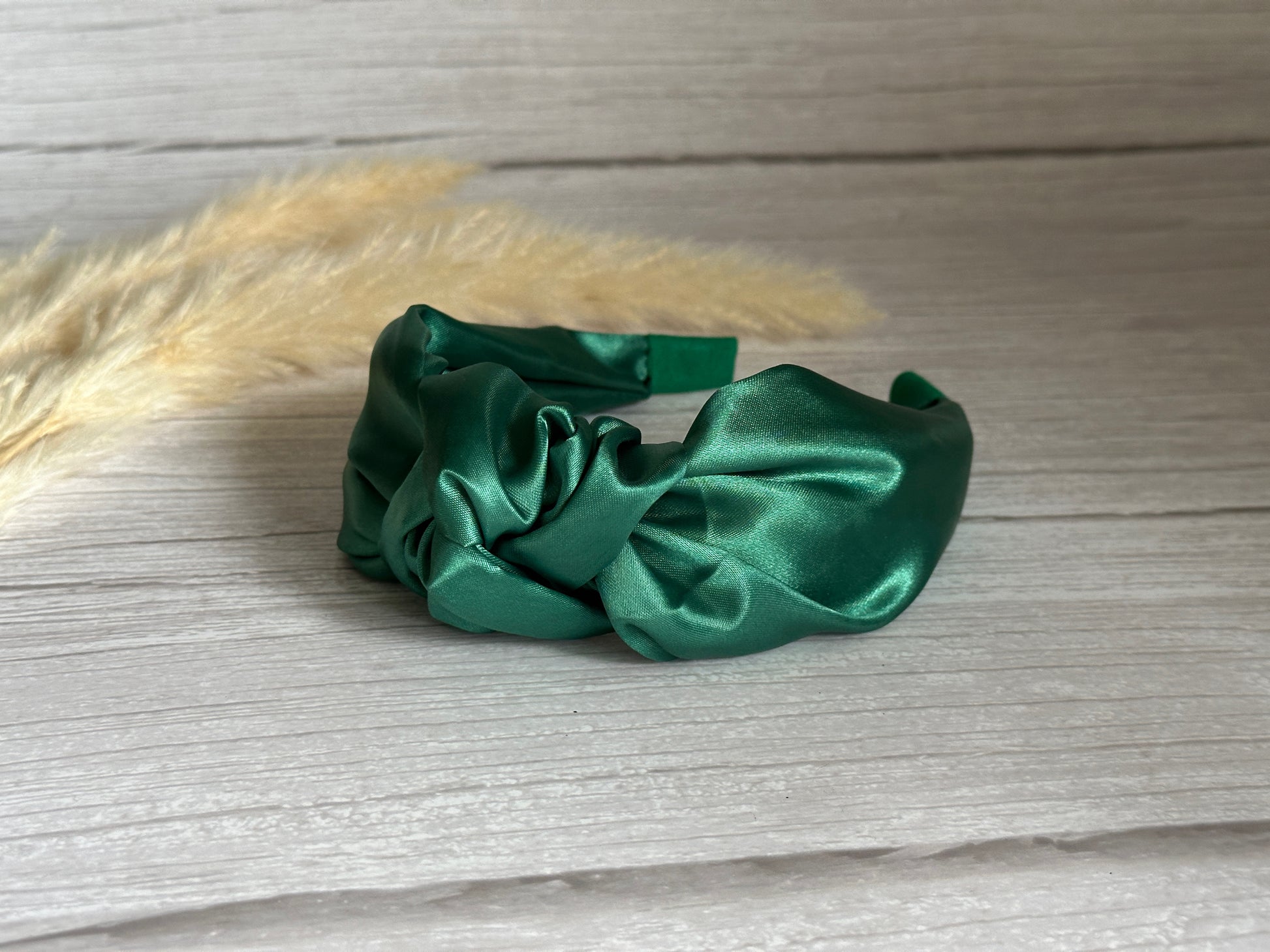 The Silk Knotted Headband in emerald green from Crafts by Kate is elegantly displayed on a light wooden surface, exuding the charm of a luxury accessory. The background is adorned with beige pampas grass, enhancing the scene with a touch of natural elegance.