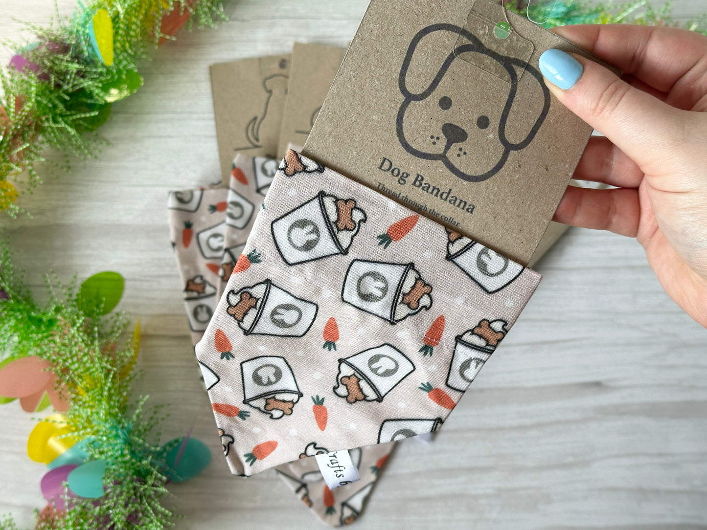 A hand displays a handcrafted cotton pet bandana by Crafts by Kate, featuring a carrot and bunny pattern for Easter Pupcup fans. The package has a cartoon dog face, while colorful flower decorations enhance the light wood background.