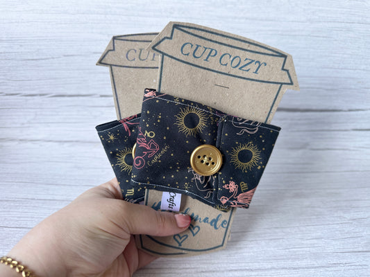 A person holds two handcrafted "Cotton Reusable Cup Cozy - Horoscope Sky" by Crafts by Kate, featuring whimsical celestial designs with a zodiac Sagittarius illustration. Each cozy includes a large golden button, all set against a light wood texture for an eco-friendly touch.