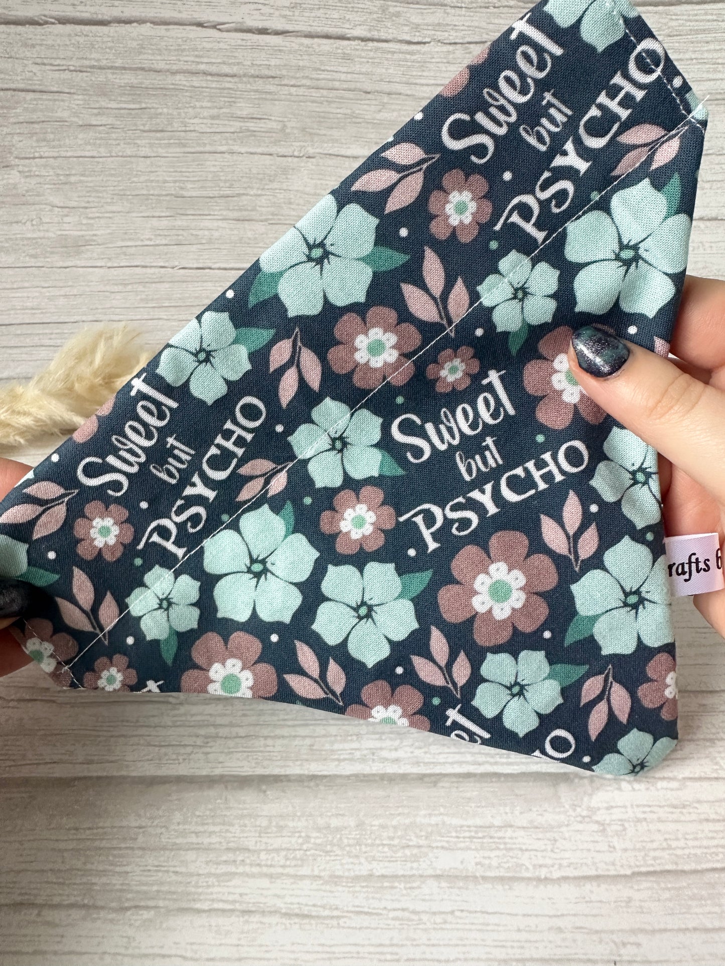 A hand holds the Crafts by Kate Cotton Pet Bandana, featuring blue, pink, and white flowers on a dark background with "Sweet but PSYCHO" in bold. This handcrafted accessory has a collar attachment and a beige rope for easy wear.