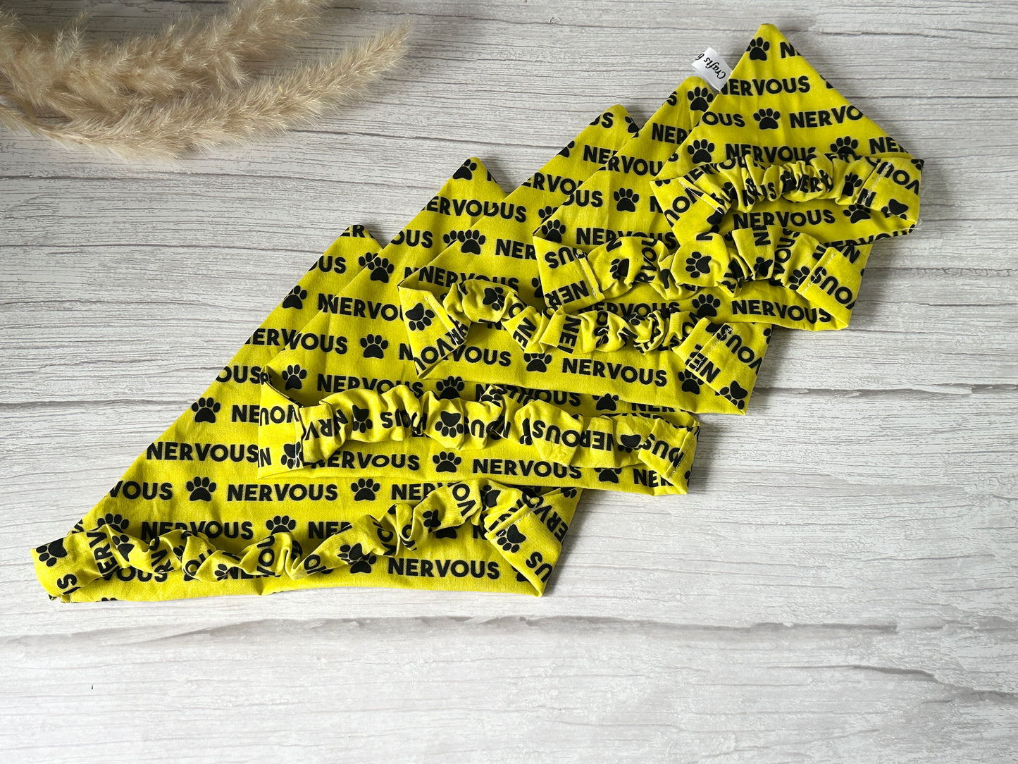 Five yellow "Cotton Pet Scrunchie Bandana - Nervous" by Crafts by Kate, featuring black paw prints and "NERVOUS" print, are fan-arranged on a light wooden surface.