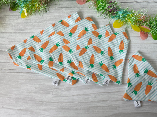 Crafts by Kate's Cotton Pet Bandanas with collar attachments, featuring striped carrots on white and blue cotton, are laid out on a wooden surface. Above them, a decorative vine of green, yellow, and pink livens up the scene.