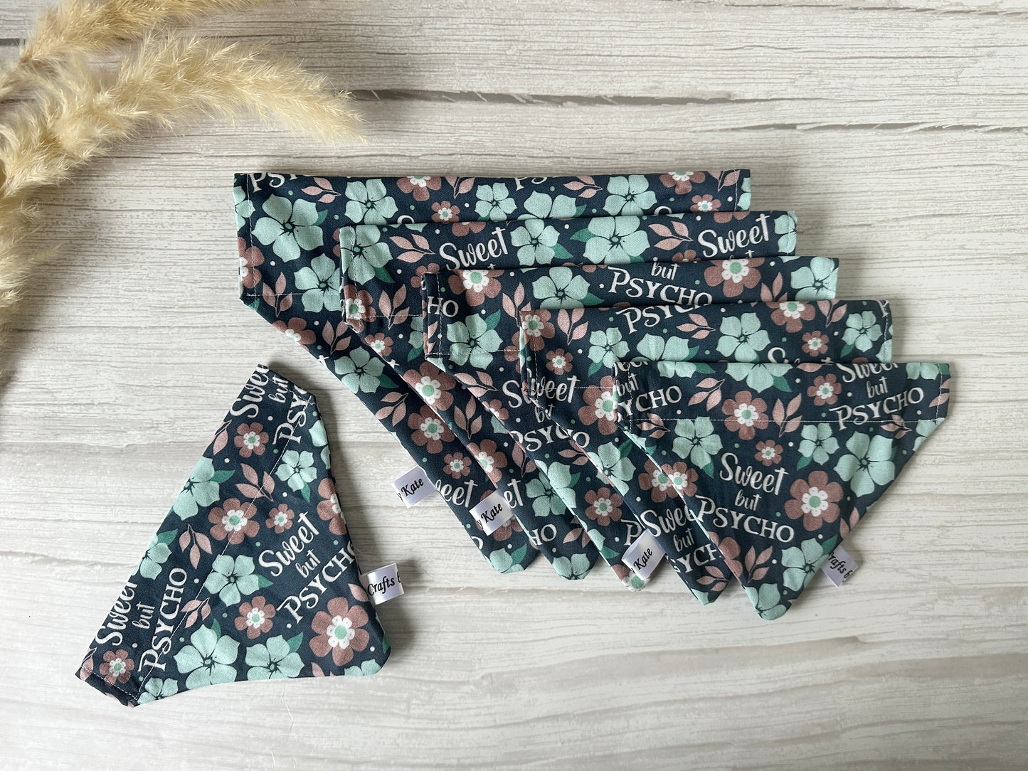 The "Sweet but Psycho" bandana by Crafts by Kate is a handcrafted cotton piece featuring mint green and pink floral patterns. Designed for collar attachment, it's displayed on light wood with dried grass in the top left corner.