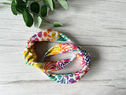 The Cotton Knotted Headband - Rainbow White Tiger Print by Crafts by Kate, with its vibrant twist design, rests on light wood near green stems—an ode to the lively spirit of the LGBTIQA+ community.
