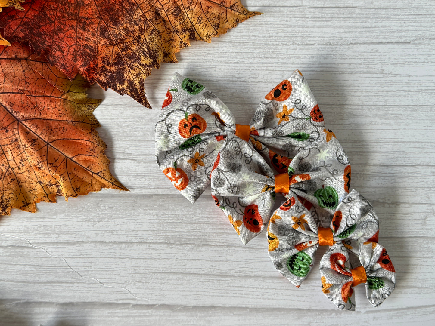 Four Crafts by Kate Cotton Pet Bows with collar attachment—featuring glow-in-the-dark pumpkins—are displayed on a white wooden surface beside autumn leaves. Ideal for pets to wear at fall festivities.