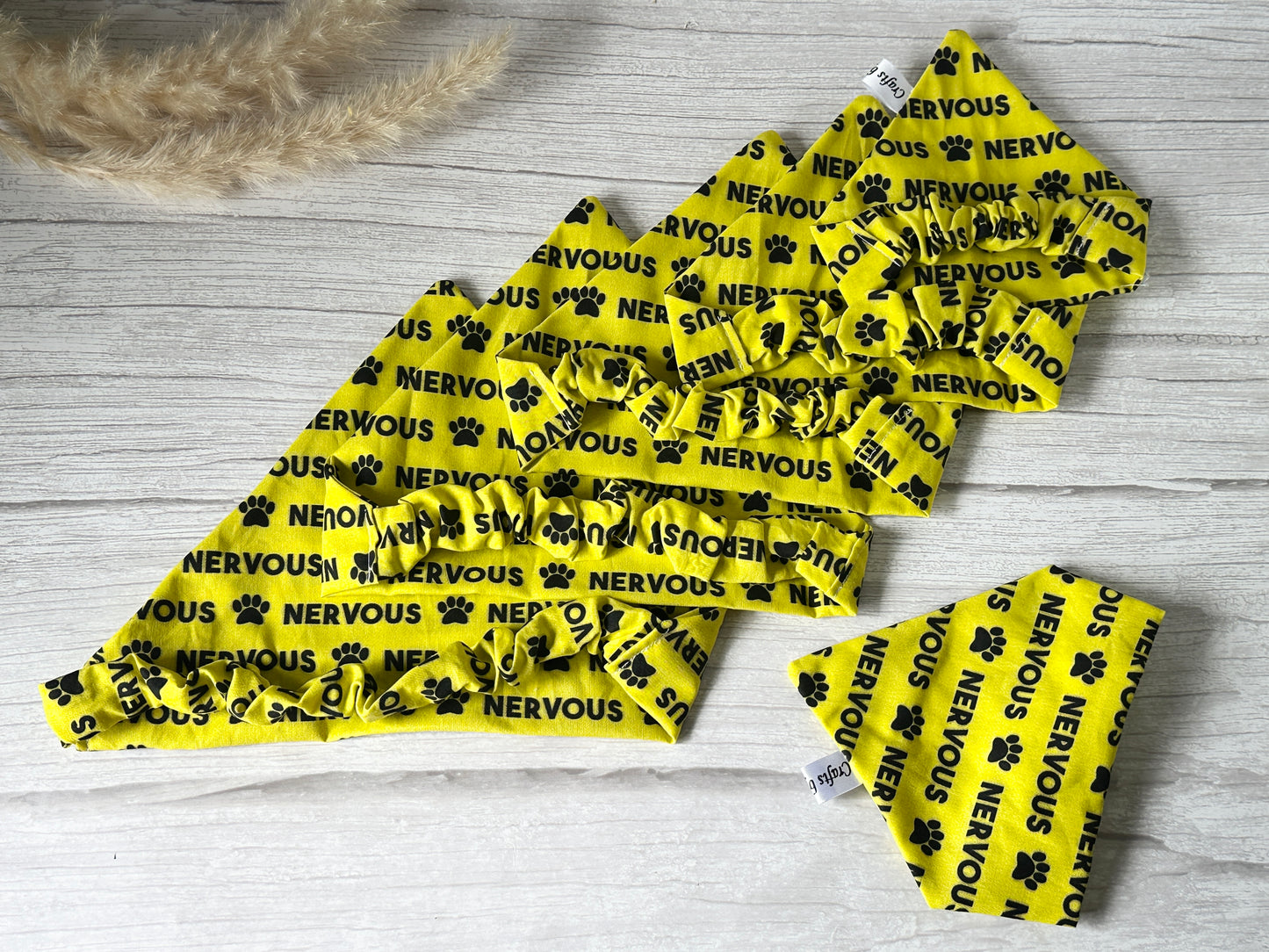 Five "Cotton Pet Scrunchie Bandana - Nervous" by Crafts by Kate, yellow with black paw prints and "NERVOUS" text, are displayed on a light wooden surface with beige pampas grass in the corner.
