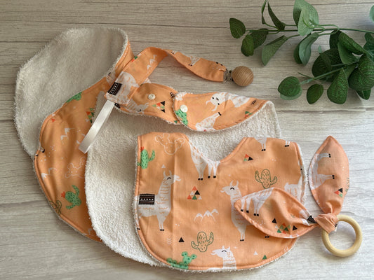 Three ultra-soft baby bibs from the "Cotton & Terry Towelling Baby Sets - Llama Loves" by Crafts by Kate, featuring llama and cactus designs, are neatly arranged on a light wooden surface with a nearby small green plant.