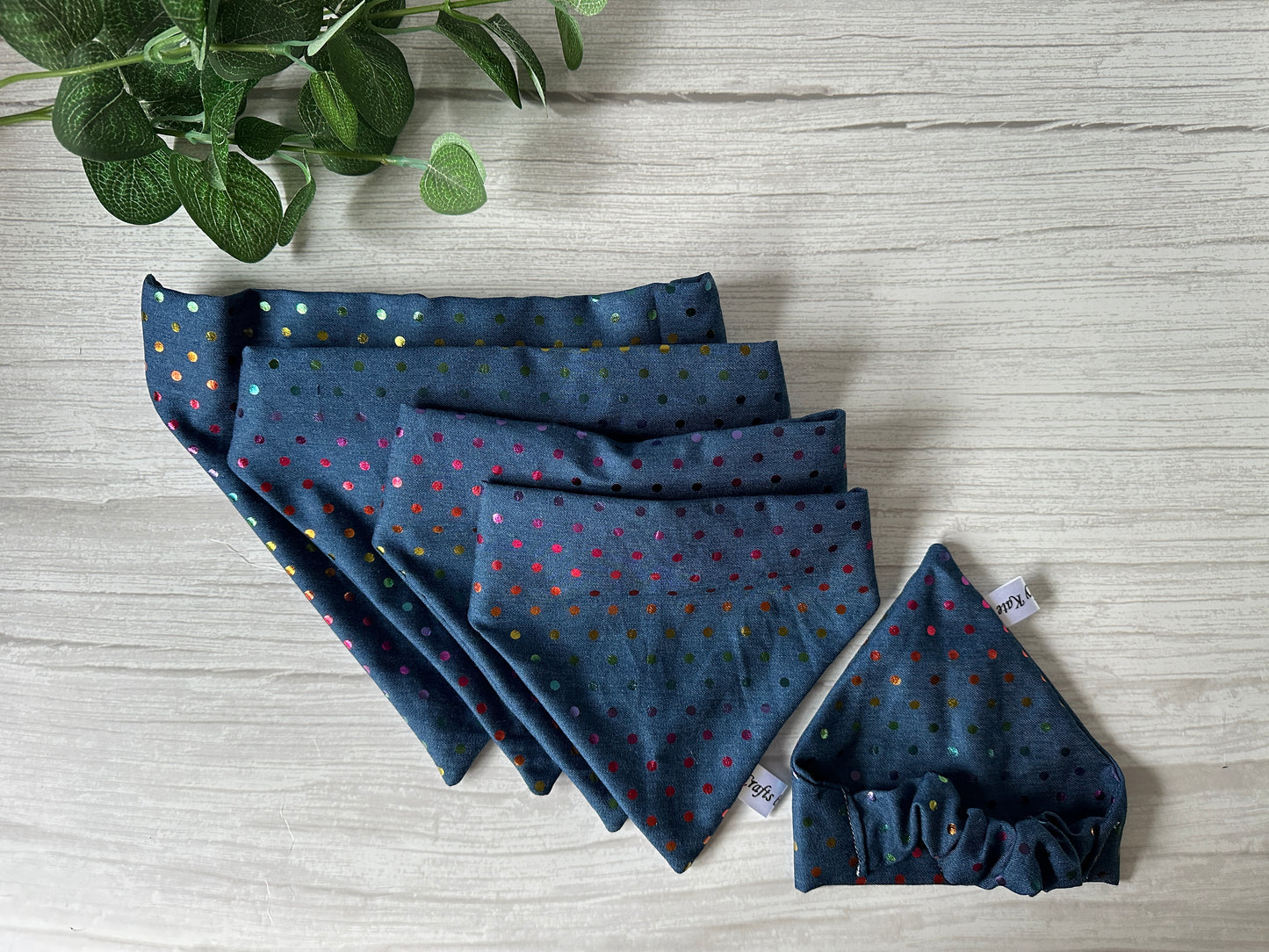 Crafts by Kate's Cotton Pet Scrunchie Bandana in Rainbow Polka Dot Foil Print is stylishly presented on a light wooden backdrop, with a leafy plant peeking in the top left corner, symbolizing LGBTIQA+ Community Support.