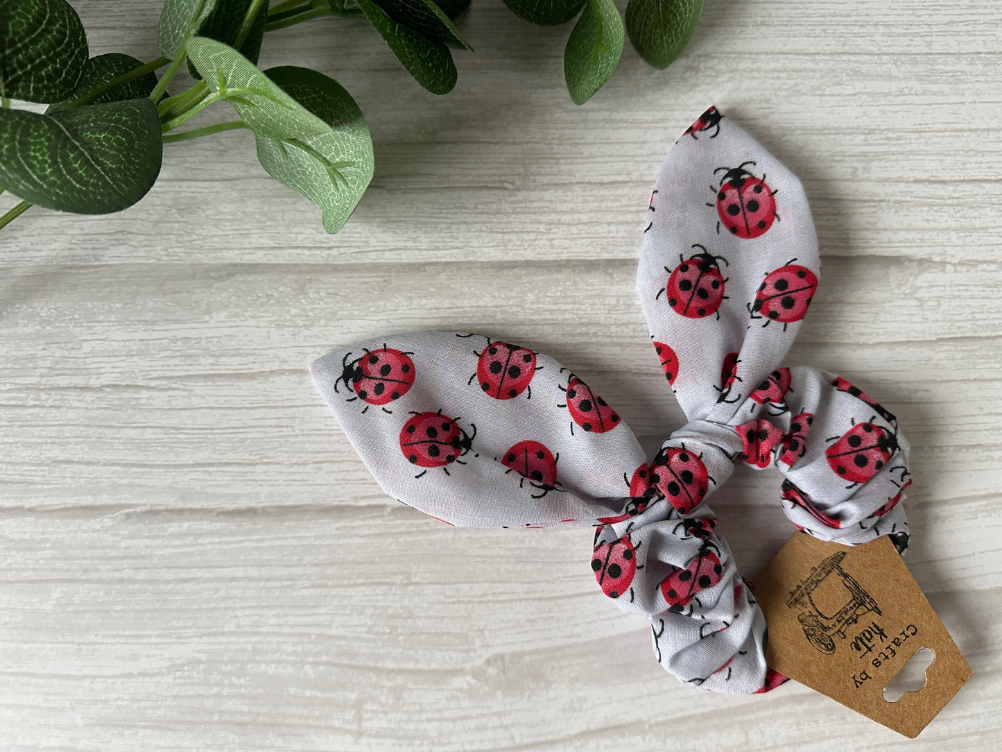 A "Cotton Scrunchie with tails - Grey Ladybirds" from Crafts by Kate sits on a light wooden surface, enhanced by green leaves that add a luxurious, natural touch to the elegant handcrafted accessory.