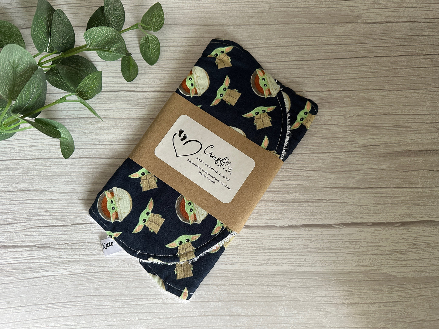 A collection of cotton and terry towelling baby burping cloths featuring a Baby Yoda design by Crafts by Kate, wrapped in brown paper labeled "Crafty Final Frontier." The cloths are displayed on a wooden surface with a green leafy plant nearby.