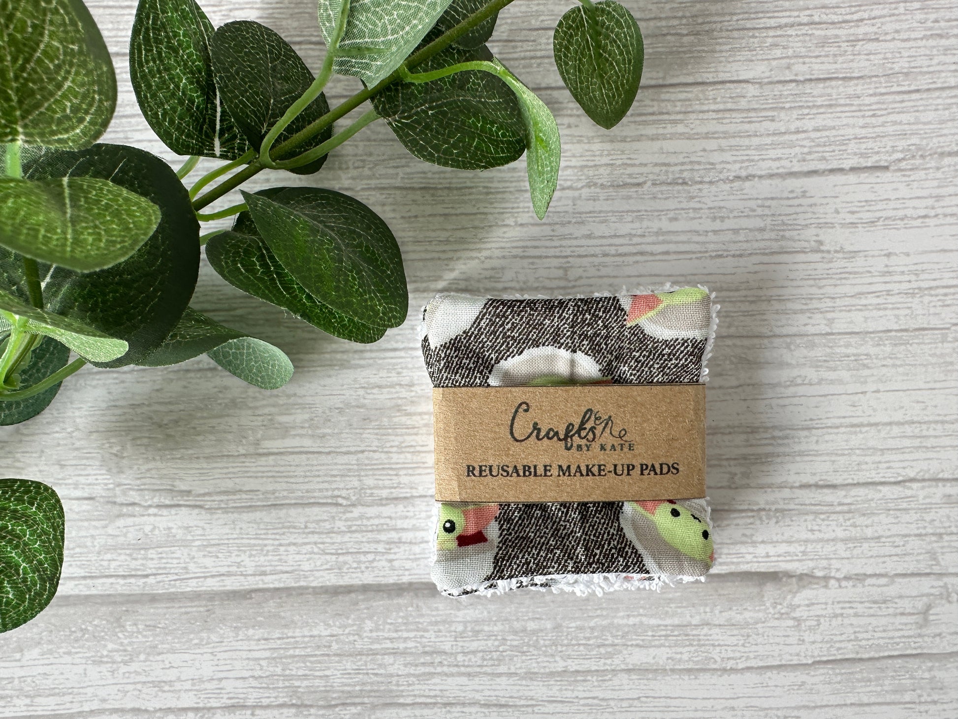 A sustainable Star Wars Grogu makeup pad by Crafts by Kate, wrapped in a brown paper label, lies on a light wooden surface with green leaves, representing the future of reusable make-up remover pads.