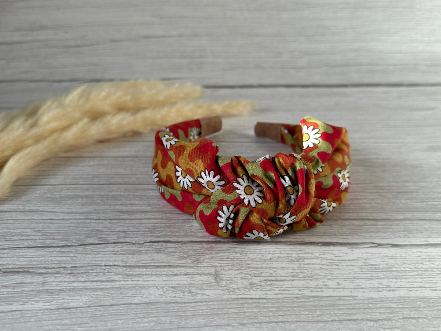 The Cotton Knotted Headband - Groovy Daisy by Crafts by Kate, featuring a red, orange, and green camo pattern with white daisies, is displayed on light wood with beige dried wheat stalks in the background.