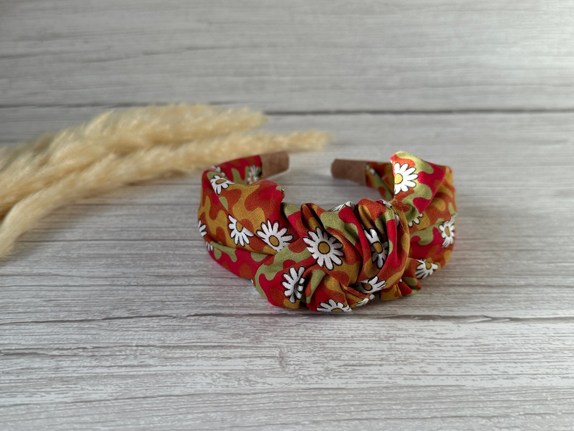 The Cotton Knotted Headband - Groovy Daisy by Crafts by Kate, featuring a red, orange, and green camo pattern with white daisies, is displayed on light wood with beige dried wheat stalks in the background.