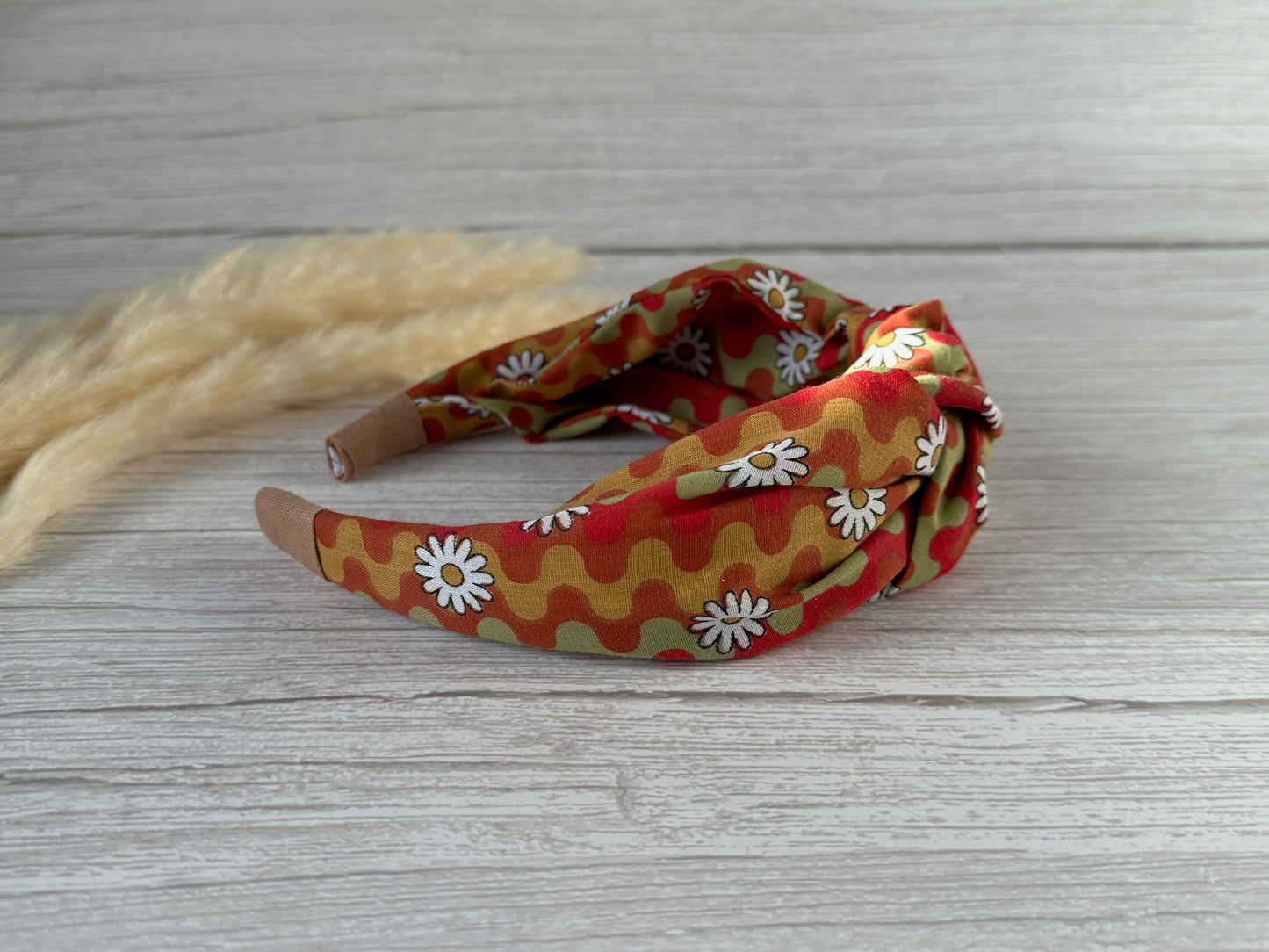 The Cotton Knotted Headband - Groovy Daisy by Crafts by Kate, featuring a wavy orange, yellow, and green pattern with white daisy flowers, rests on a light wooden surface beside beige dried grasses.