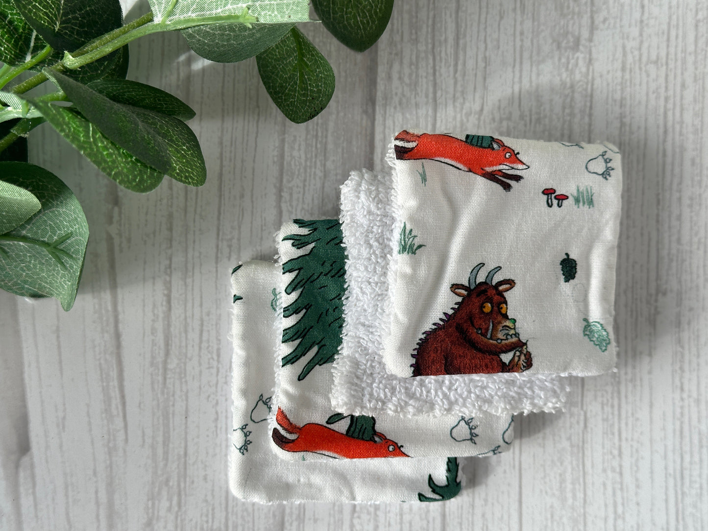 Three white, handcrafted Cotton Reusable Make-up Remover Pads - Gruffalo in the Forest by Crafts by Kate are stacked on a light wood surface. Each pad features different scenes from the children's book, "The Gruffalo," with characters like the orange fox running and the Gruffalo holding a nut. Green leaves decorate the left corner.