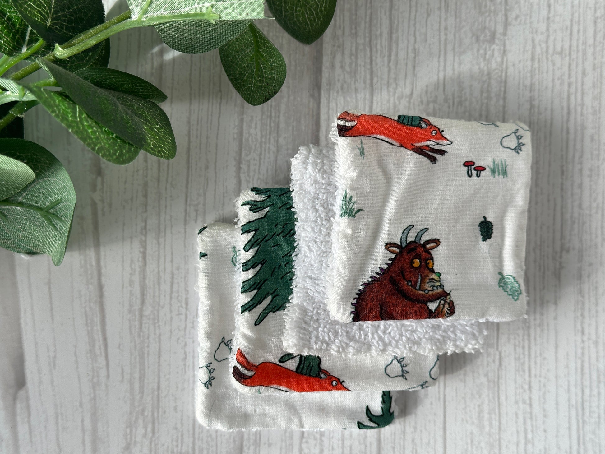 Three white, handcrafted Cotton Reusable Make-up Remover Pads - Gruffalo in the Forest by Crafts by Kate are stacked on a light wood surface. Each pad features different scenes from the children's book, "The Gruffalo," with characters like the orange fox running and the Gruffalo holding a nut. Green leaves decorate the left corner.