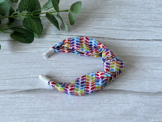The Cotton Knotted Headband - Rainbow Leaves by Crafts by Kate features luxury cotton with a twisted knot and rainbow leaf print, shown on light wood with green leaves nearby.