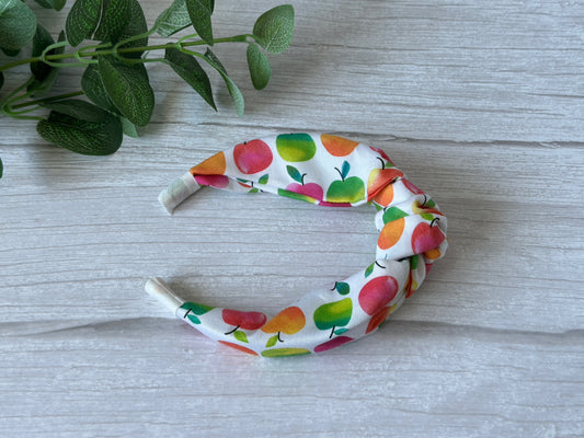 The Crafts by Kate Cotton Knotted Headband - Rainbow Apples, featuring colorful apple patterns on white cotton, rests on a light wood surface with green leafy plant stems in the upper left corner.