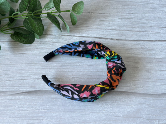 The Cotton Knotted Headband - Rainbow Tiger Print by Crafts by Kate sits atop a light wood surface, with green leafy plant stems in the upper left corner.