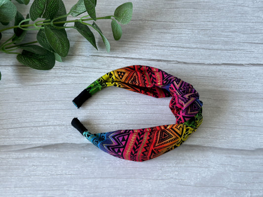 The Cotton Knotted Headband - Rainbow Aztec Print by Crafts by Kate is displayed on a light wood surface beside green leafy plant stems.